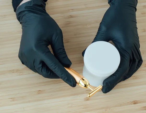 Black-gloved hands holding a gold applicator beside a white treatment jar on a wooden table