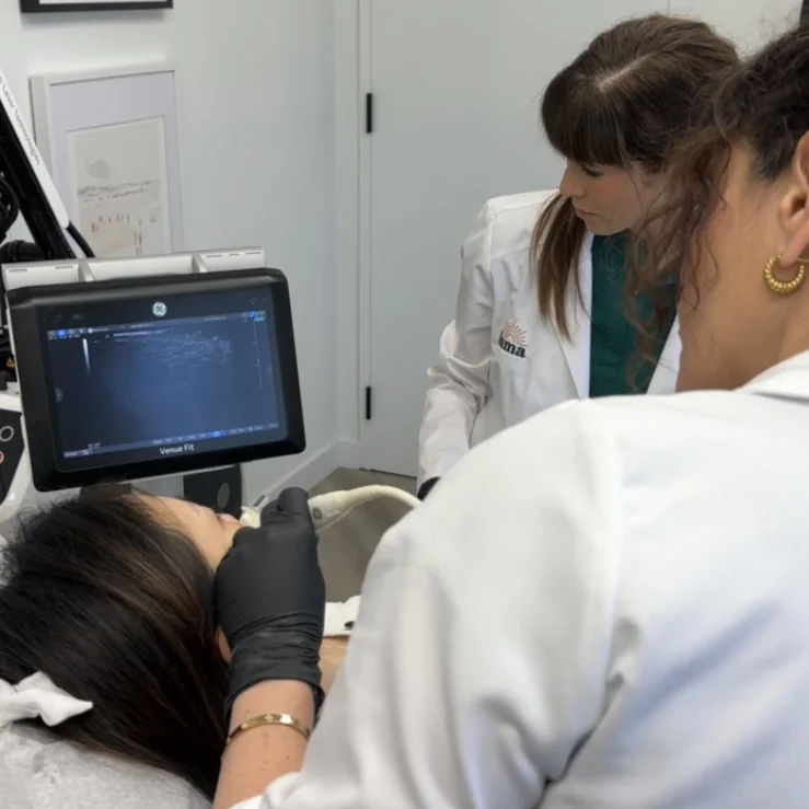 A GE Venue Fit ultrasound monitor showing a live scan while a patient is scanned and a second practitioner in a luma coat observes